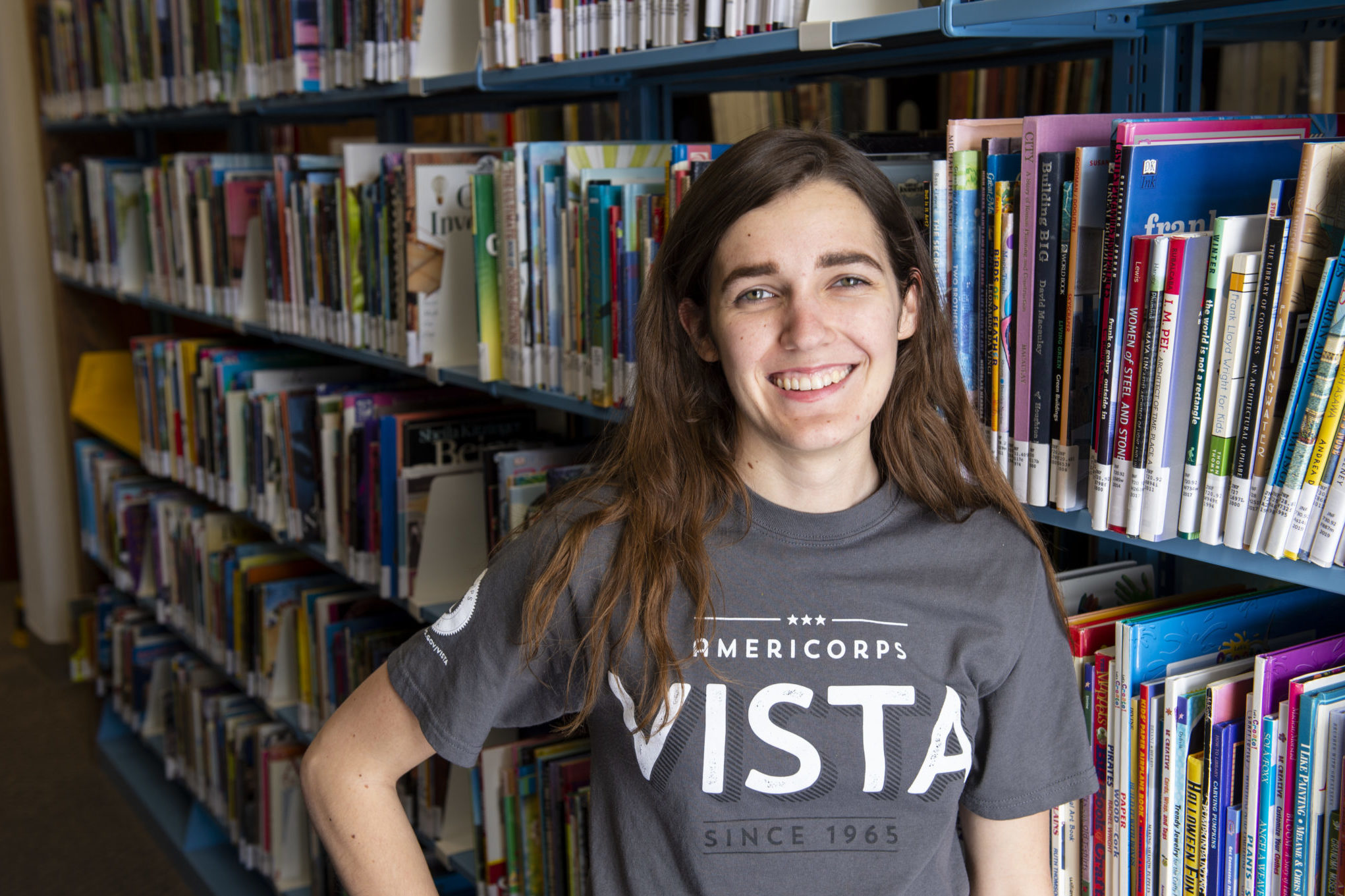 woman standing in front of books in a library wearing a grey shirt that says VISTA
