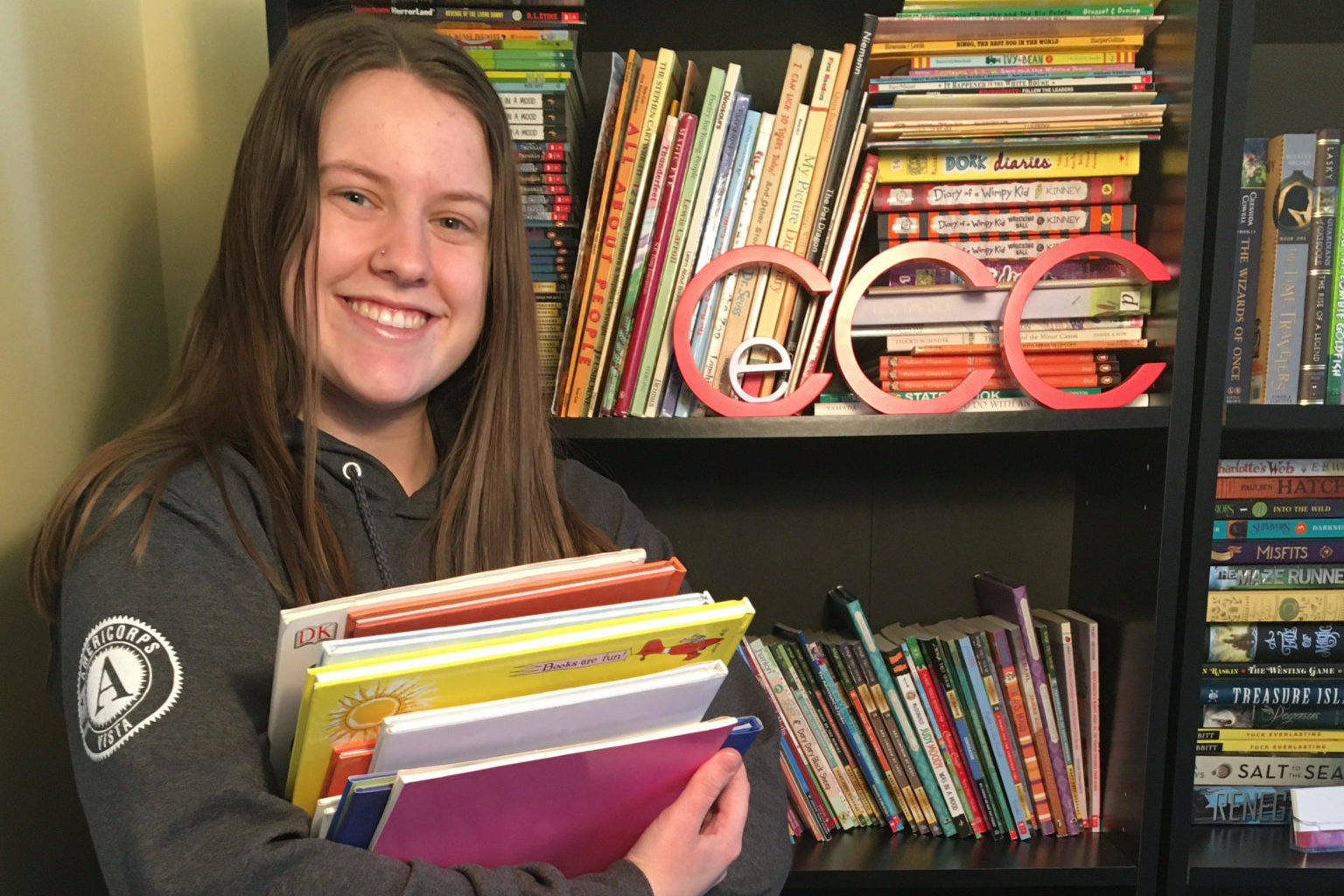 woman wearing a VISTA sweatshirt standing in front of a book shelf holding a stack of books