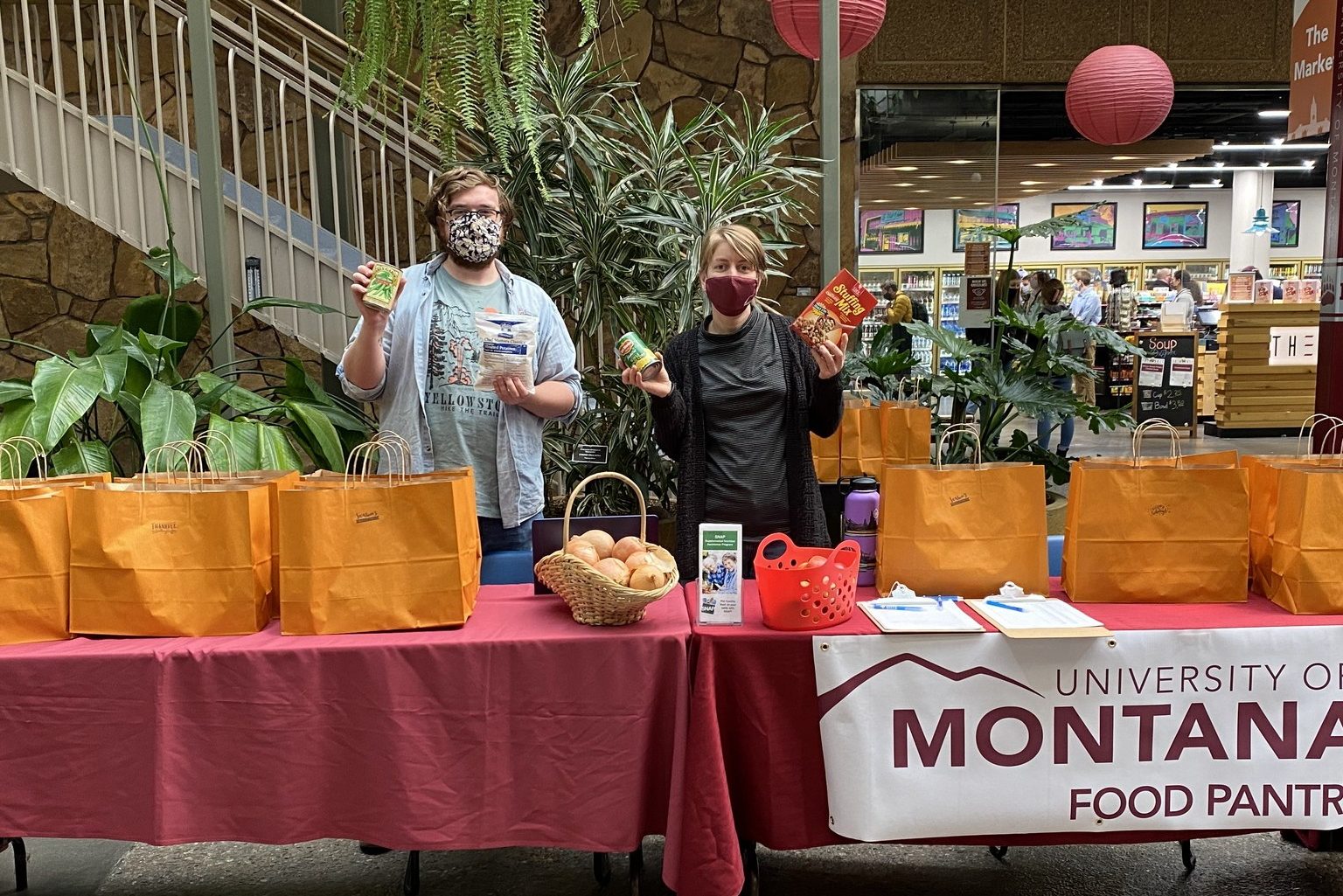 two people who are standing behind a table for the University of Montana Food Pantry while holding food