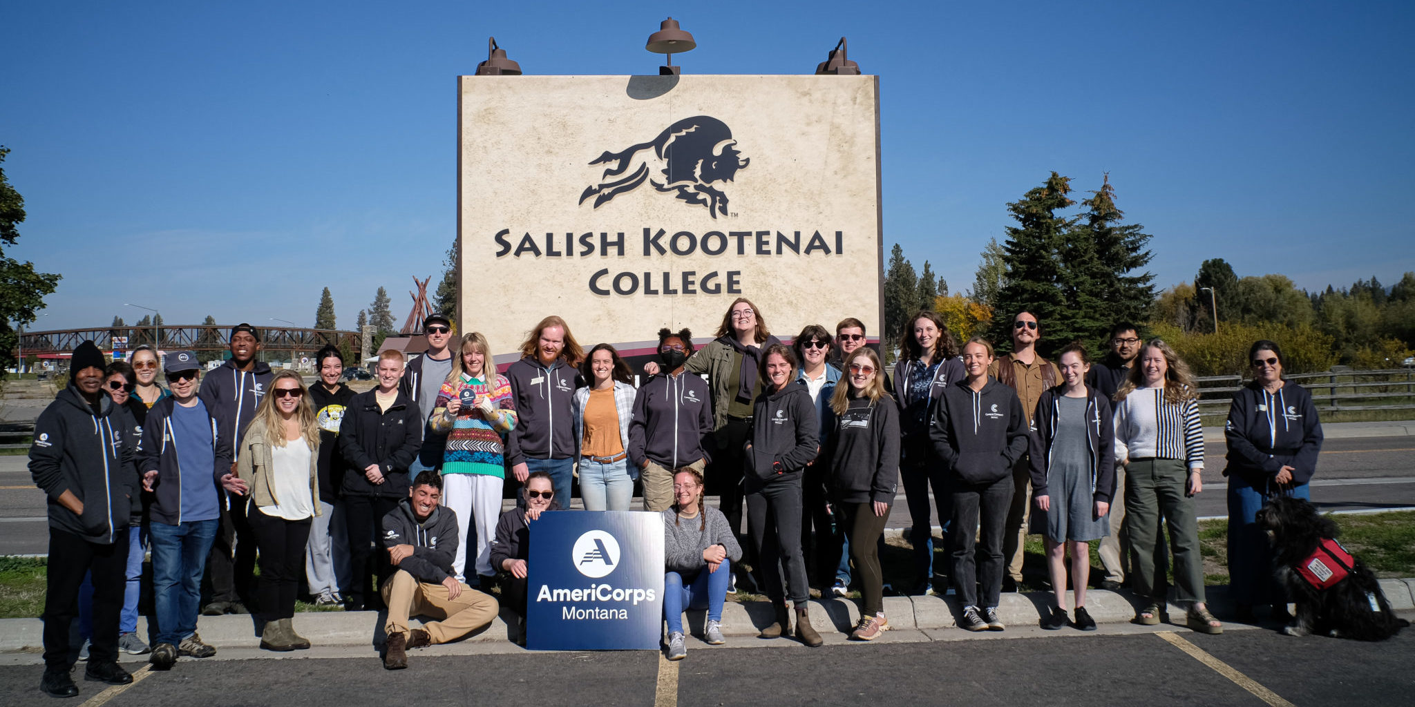 Group photo af about 40 AmeriCorps members and MTCC staff. They are holding a sign with the AmeriCorps logo, and there is a large sign that says "Salish Kootenai College" behind them. On the sign is a stylized image of a charging buffalo.