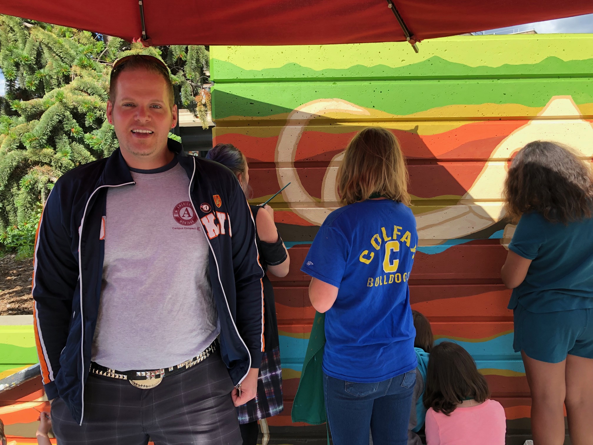 An AmeriCorps member smiling at the camera while students paint a colorful mural of a mammoth fossil in the background.