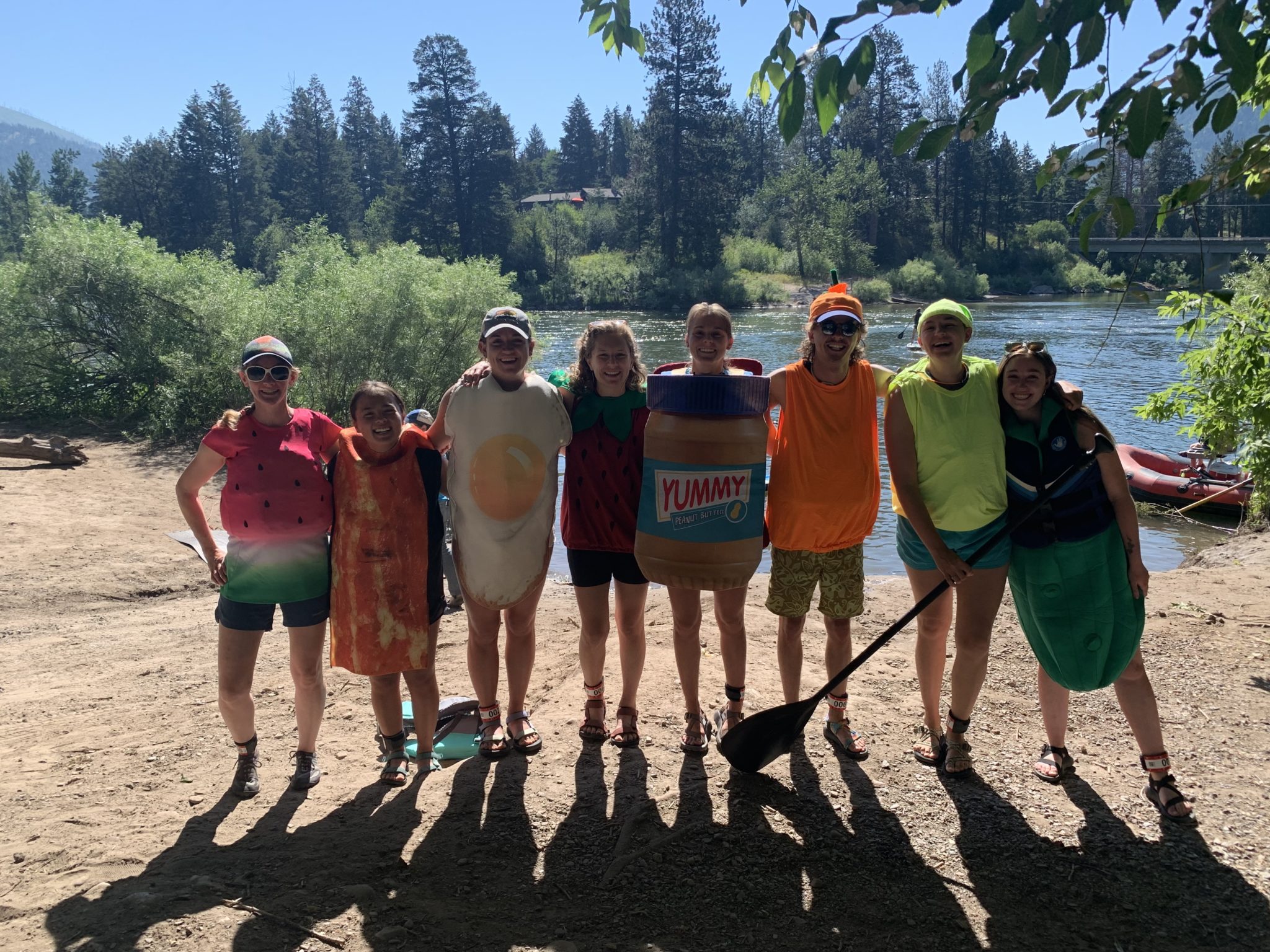 A group of eight people on a riverbank. Many of them are dressed in silly food costumes: watermelon, bacon, fried egg, strawberry, peanut mbutter, and pickle.