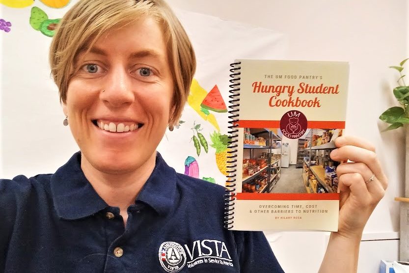 AmeriCorps member holding a "Hungry Student Cookbook" and smiling at the camera.